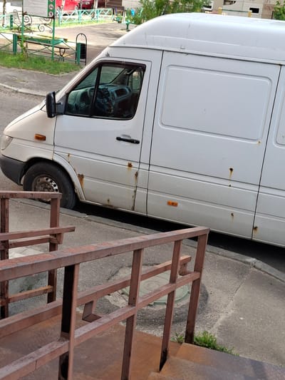 The image shows the side of a white cargo van parked on an asphalt surface. The van has visible rust spots and some dents. A metal staircase, appearing to be made of rusted metal bars, is positioned to the right of the van, partially obscuring the side of the vehicle. The staircase is angled, suggesting it might be a temporary structure or part of a larger construction.
In the background, a building with multiple windows is visible, along with some trees and greenery. A green metal fence with decorative elements and some signage is also present. The overall scene appears to be an outdoor urban or semi-urban environment, possibly a residential area or a service area. The lighting suggests it is daytime, with no specific indication of the time of day or weather conditions. There is no visible activity or people in the frame. The crops provide closer views of the van's body, the staircase, and the surrounding environment.