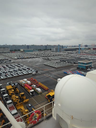 The image shows a vast outdoor car storage lot in the city of Yokohama, Japan, under a cloudy sky. The lot is filled with rows upon rows of white and dark-colored cars, neatly parked. In the foreground, a large white dome-shaped object, likely part of a ship or a satellite dish, occupies a significant portion of the frame. Next to it, a red life preserver is visible, attached to a railing. Several construction vehicles, including yellow excavators and loaders, are parked in a separate area. In the background, a multi-story building complex and various industrial structures, including cranes and shipping containers, are visible, suggesting a port or industrial area. The ground is wet, indicating recent rain. The scene is devoid of people.