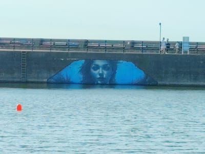 The image shows a mural of a woman's face on the side of a sea wall in Wallasey, United Kingdom. The mural is predominantly blue and depicts the woman's face with dark hair, large eyes, and prominent lips. The sea wall is a concrete structure with a darker, rougher texture, and a ladder is visible on its left side. Above the sea wall, a promenade is visible with benches and a railing. Three people are standing on the promenade, two of whom appear to be walking. The sea in the foreground is a light blue with ripples, and a single orange buoy is floating in the water. The sky is a pale blue and appears hazy.