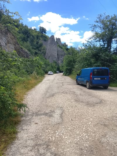 The image depicts a dirt road winding through a natural landscape with steep rocky cliffs on the left and dense green trees on both sides. Several cars are visible on the road, including a blue van in the foreground. The sky is blue with scattered white clouds. The scene appears to be in a rural or natural area, possibly a park or nature reserve, located in the vicinity of Asparuhovo, Bulgaria. The overall impression is one of a scenic drive or an outdoor excursion.