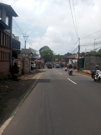 The image displays an active two-lane asphalt road in Niz Lakuk, India, under a bright, overcast sky. The road features a faded white centerline and a solid white line delineating the left shoulder. On the immediate left, a dirt shoulder borders a low concrete fence, behind which are multi-story residential or commercial buildings. Numerous utility poles with extensive, tangled overhead wiring are prominent on both sides of the road, particularly dense on the left.

Traffic is active, with vehicles moving in both directions. Approximately seven vehicles are clearly visible. In the foreground on the right, a person wearing a blue jacket and a helmet is riding a light-colored scooter, moving away from the viewer. Further down the right lane, two individuals on dark scooters, also wearing helmets and dark clothing, are traveling towards the viewer. A black SUV and a blue van are visible in the left lane, moving away from the viewer, with additional cars and scooters in the distant traffic.

Along the left side of the road, past the fence, a small green shrub and various commercial or residential structures with unreadable signage are present, including a white SUV partially parked off the road. On the right side, basic structures, stacked wooden planks, and clothes hanging outside a building are visible near a utility pole. The background features dense green foliage and trees covering elevated terrain or hills, extending along both sides of the road as it curves slightly to the left in the distance.