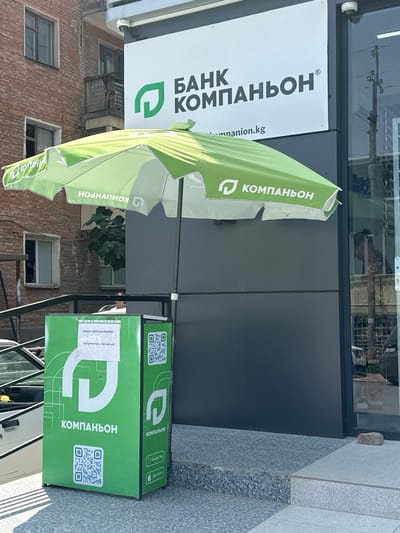 The image shows the exterior of a "Bank Kompanion" branch in Kadamjay, Kyrgyzstan. A green promotional umbrella, also branded with "Kompanion", is set up in front of the bank. Below the umbrella is a green promotional box with the "Kompanion" logo and QR codes. A white sign is affixed to the box. The bank building has a modern facade with large windows. In the background, a brick building with balconies is visible. The overall scene suggests a street-level bank branch with an outdoor promotional display.