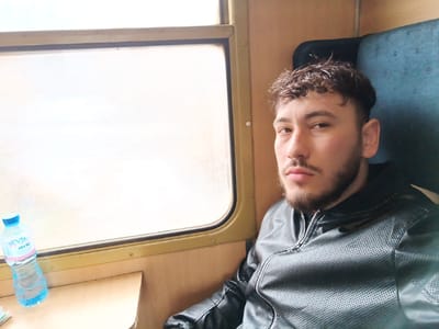 The image depicts a male individual seated within the interior of a passenger vehicle, identified as being in Старо село, Bulgaria. The subject is a male with medium skin tone, dark, curly hair, and a short beard and mustache. He is wearing a dark, possibly black, perforated synthetic leather jacket with a metallic zipper. He looks directly forward, facing the camera, with a neutral facial expression.
To the left of the subject, a rectangular window with rounded corners reveals an overexposed white exterior, suggesting bright light conditions or motion outside. Below this window, a light-colored wooden or laminate surface serves as a small table. On this surface, a clear plastic bottle of water with a blue cap and a light blue label bearing the Cyrillic text "ДЕВИН" (Devin) is positioned. The interior walls of the compartment feature light-colored wood-grain or beige laminate paneling. The subject is seated against a dark blue fabric seat with a dark headrest visible behind him. The compartment is uniformly lit, with a primary light source originating from the window.