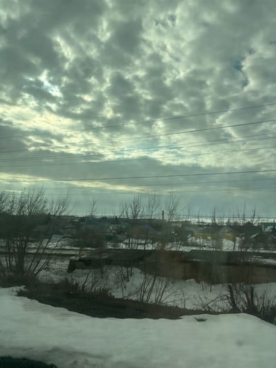 The image presents an elevated view of a winter or early spring landscape in Nurlat, Russia, under a heavily overcast sky featuring textured, grey-white clouds with brighter patches indicating obscured sunlight in the upper left. Multiple dark, horizontal utility lines traverse the upper-mid section of the frame. Below these lines, the distant horizon reveals a flat, snow-covered expanse, possibly a frozen body of water or field, under a lighter sky. The midground contains a sparse residential settlement with various small buildings, identifiable by their roofs in shades of dark green, dark red, brown, and grey. Bare-branched deciduous trees and dense shrubs are scattered throughout the settlement and in the foreground. A singular, slender, vertical structure, possibly a chimney or vent, is visible above the houses. The ground is largely covered in snow, with areas of dark, exposed earth and vegetation, particularly prominent in the foreground. No people, specific actions, or interactions are visible within the scene.