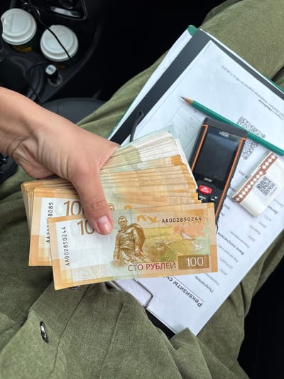 A hand holds a thick stack of Belarusian 100 Ruble banknotes. The banknotes are predominantly light orange and beige, with images of historical figures and national symbols. One banknote clearly displays "100" and "СТО РУБЛЕЙ" (100 Rubles) in large font, along with a serial number "AA002850244" and the year "2022". Another visible serial number is "AA018085". The background includes a document with Russian text, a pencil, a handheld measurement device labeled "RGK D120", and two white paper cups with lids, possibly containing beverages. The overall setting appears to be inside a vehicle, with a portion of a green jacket visible in the foreground. The document in the background contains Cyrillic text including "ЛИЦЕНЗИЯ № 1481 от 11.08.2015", "Сбербанк", and other financial transaction related terms. The date "11.08.2015" suggests the document is from a Russian bank. The image has no visible connection to the city of Sedanovo, Russia.