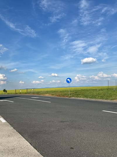 The image shows a road in Cruceni, Romania. The road is made of asphalt and has white lane markings. Next to the road is a field of green grass, enclosed by a fence. There is a blue road sign with a white arrow indicating a turn to the left. The sky is blue with scattered white clouds.
