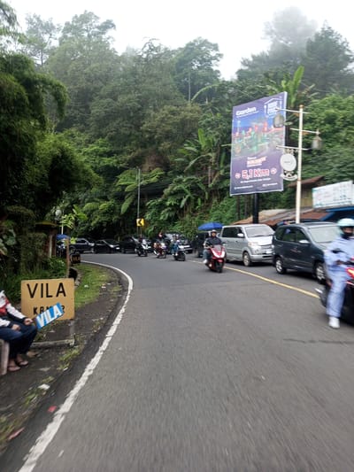 The image depicts a road scene in a tropical or subtropical environment. A paved road curves to the left, with a white line marking the edge. On the left side of the road, a person sits next to a sign that reads "VILA". The person is wearing a casual outfit consisting of what looks like a jacket and pants, and flip-flops on their feet. Behind the person, there is lush green vegetation, including bamboo.
Vehicles, including cars and motorbikes, occupy the road. There is an advertisement billboard on the right side of the road advertising a "Garden". The surrounding environment is marked by dense trees and foliage, with fog or low-lying clouds in the distance.