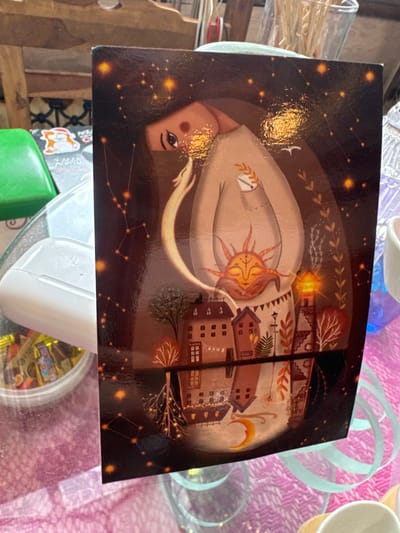 A glossy print or postcard is centrally positioned on a transparent tabletop in an indoor setting in Matveevskoe, Russia. The print features a stylized artistic depiction: a light-skinned female figure with long dark hair and dark eyes, marked with a small red circle on her right cheek, occupies the upper portion. She wears a light-colored, long-sleeved garment with a sprig of wheat in a visible pocket on her left chest. Her hands cradle a stylized sun motif with a closed-eye face. A pale, crescent-shaped object originates from her left chest area and flows downwards along her side. The background is a dark, starry sky with visible constellations, including what appears to be Taurus to the left. Small white bird silhouettes are present in the upper right. Below the figure, a cityscape is depicted, featuring several multi-story buildings with brown and reddish roofs, some exhibiting illuminated windows. Autumnal trees and a glowing lighthouse are also part of this urban landscape, with a string of banners connecting buildings. The lower part of the print shows a reflection or inverted representation of the cityscape, featuring roots and glowing lights, with a yellow crescent moon visible.
Surrounding the print, various objects are visible on the transparent table surface. To the left of the print, a bright green plastic lid, a white elongated object (resembling an earbud case), and a clear round container filled with individually wrapped candies are present. Beneath the transparent table, a pink or light purple patterned fabric or tablecloth is visible. To the right of the print, a clear glass vessel containing dry, light brown plant material (possibly straw or twigs) is visible. A white ceramic mug with a light brown wooden handle is partially visible at the bottom right, and a light blue ribbon or string is draped on the tablecloth. Blurred wooden elements are discernible in the background, suggesting furniture.