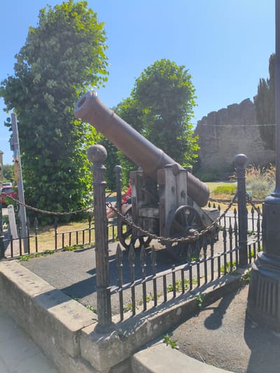 The image depicts a historic cannon, likely a relic from Ludlow Castle, positioned outdoors in what appears to be a public space within Ludlow, United Kingdom. The cannon rests on a wooden carriage with two large wheels. It is secured by a metal chain and is behind a decorative black metal fence with pointed finials. A stone wall, characteristic of historical fortifications, is visible in the background, as are lush green trees and a clear blue sky. The cannon is situated on a paved area with a low stone border in the foreground. A portion of a black lamppost is visible on the right side of the frame. Sunlight casts shadows on the ground, indicating it is a bright day. A person wearing a red shirt is seated on the carriage of the cannon.