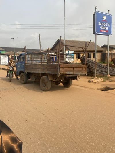The image captures a street scene in Igbesa, Nigeria, under an overcast sky. A blue, six-wheeled flatbed truck with rusty metal cage-like sides is stationary on a light brown dirt road, facing away from the camera. The truck displays "FUZE" or similar text on its cab.
On the left side of the truck, a green motorcycle is stopped. An individual, wearing camouflage-patterned clothing and blue trousers, is seated on the motorcycle, facing towards the truck's driver side.
To the right of the truck, a prominent blue rectangular sign is erected on two metal poles, displaying "DANGOTE CEMENT" with a right-pointing arrow and the Nigerian coat of arms. Adjacent to this sign, a substantial pile of rebar (reinforcing steel bars) is stacked on the ground. A square, open drainage culvert is visible in the dirt beside the road.
In the background, several single-story buildings with various roof types are present. One building features a partially visible white sign with Arabic script and English text. Multiple utility poles, including one with a solar panel, and overhead power lines traverse the frame. The immediate foreground on the left shows a portion of dark, patterned clothing.