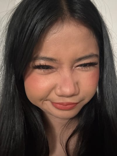 A close-up, head-and-shoulders shot captures a young woman of Asian descent with long, straight, black hair against a plain, light-colored background. She is looking directly at the camera with a playful, squinting expression and a slight smirk. Her eyes are lined with dark eyeliner and she is wearing false eyelashes. Her cheeks are flushed with a rosy blush, and her lips are painted with a coral-toned lipstick. The lighting is bright and evenly distributed, highlighting her facial features and the texture of her skin. The image appears to be a selfie taken indoors, likely in a residential setting in North Jakarta, Indonesia.