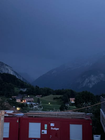This is an outdoor, eye-level, medium shot photograph taken at dusk or early evening in Hasliberg Reuti, Switzerland. The foreground is dominated by two large, red, modular shipping containers, stacked side-by-side. The left container has a white-framed window. The right container displays the logo "Frutiger" in white lettering, along with a white roller shutter door. Stacks of lumber and wooden planks are piled on top of the containers. An orange cord stretches across the scene from the right side of the frame, attached to a fence-like structure.

In the mid-ground, a small village is nestled on a green hillside. Numerous houses with lit windows are visible, indicating human habitation. Trees and foliage are scattered throughout the village and surrounding hills.

The background features a dramatic mountain landscape. The mountains are dark and shadowed, with some exposed rock faces that are lighter in color. The sky above is a deep, dark blue, transitioning to a lighter blue near the horizon, suggesting the last vestiges of daylight. The overall atmosphere is serene and twilight. There are no people or animals visible in the image.
