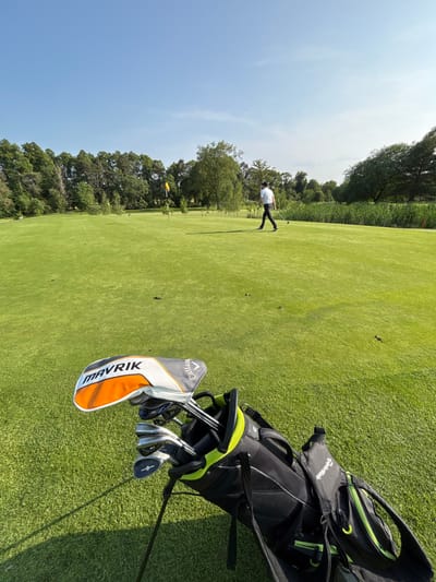 A golf bag, with a black and green exterior, sits on a green golf course. The bag contains a set of golf clubs, including a driver with an orange "MAVRIK" logo. A man in a white shirt and black pants walks away from the camera in the distance, walking across the course. The scene is located on a golf course in Pocklington, United Kingdom, at latitude 53.935 and longitude -0.754. There is a bright blue sky in the background and trees can be seen in the distance. The ground is covered in green grass.