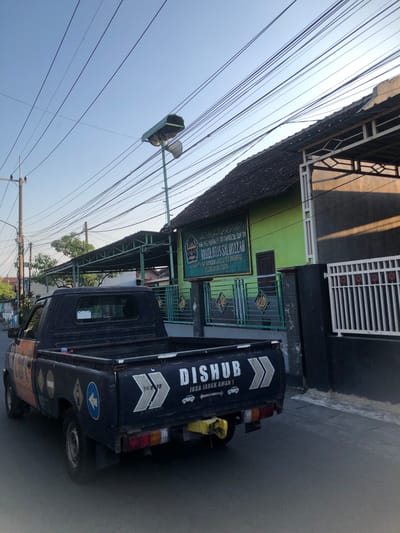 In Kalidawir, Indonesia, a dark blue pickup truck with orange accents on its side is parked on a paved road. The truck has several circular stickers on its rear panel, including one depicting a left-pointing arrow within a blue circle. The text "DISHUB JAGA JARAK AMAN!" is visible on the truck's tailgate, along with a graphic of two chevrons. A yellow hitch is attached to the rear bumper.
Behind the truck, a green building with a dark tiled roof is visible. A sign on the building reads "RAUDHATUL SALIKIN," with smaller text above and below. The building is bordered by a dark fence with white vertical bars. To the left of the truck, a utility pole with multiple wires extends upwards into a clear sky. The scene is illuminated by sunlight, casting shadows on the ground and buildings.