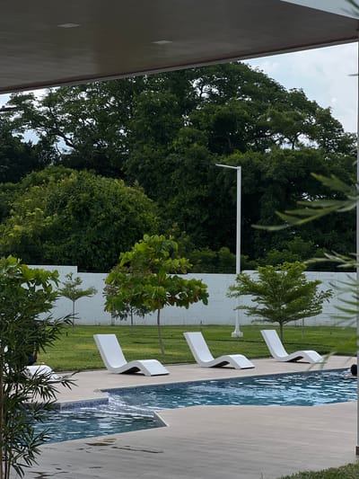 The image depicts a serene poolside scene in San Miguel, El Salvador. The primary subjects are several modern white lounge chairs arranged on a light-colored tiled patio beside a glistening blue swimming pool. The pool features a mosaic tile border and the water is rippled, suggesting gentle movement, possibly from a small waterfall or fountain as indicated by a stream of water visible entering the pool.
The setting is lush and green, with a well-maintained lawn bordering the patio and pool area. Behind the lawn, a dense line of mature, vibrant green trees forms a natural backdrop, creating a sense of privacy and tranquility. A white, textured wall, possibly a partition or a building facade, is visible in sections behind the lounge chairs and grass.
The sky above is partly cloudy, suggesting it is daytime, though the specific time is not discernible. The overall mood of the image is peaceful and relaxing, inviting one to unwind by the pool. The clean lines of the lounge chairs contrast with the organic shapes of the trees, creating a harmonious blend of modern design and nature. There is no visible text in the image.