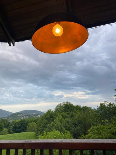 The image shows an outdoor scene under a cloudy sky, likely taken from a porch or deck. A large, round, orange-colored light fixture hangs prominently on the left side, illuminating the area. In the background, there is a green landscape with trees, grass, and hills. In the distant background, there are some buildings, likely part of a town or city. The scene appears to be in a rural or suburban area, not necessarily limited to Progres, Russia. The image does not depict any people or specific actions.