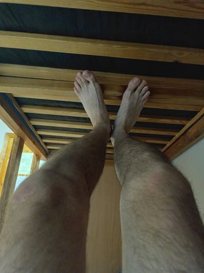 The image shows a first-person perspective of a person's legs and feet, viewed from underneath a wooden bed frame. The bed frame consists of parallel wooden slats with black fabric between them. The person's feet are bare and resting on some of the wooden slats. The legs are hairy and tanned. The background is blurry but appears to be indoors, possibly a bedroom, with a light-colored wall and a wooden door frame visible. There is a document or drawing with lines and text attached to the side of the bed frame. The lighting suggests an indoor setting, possibly during the day. The image does not contain any information that would allow for its location to be determined in the city of Kogalym, Russia.