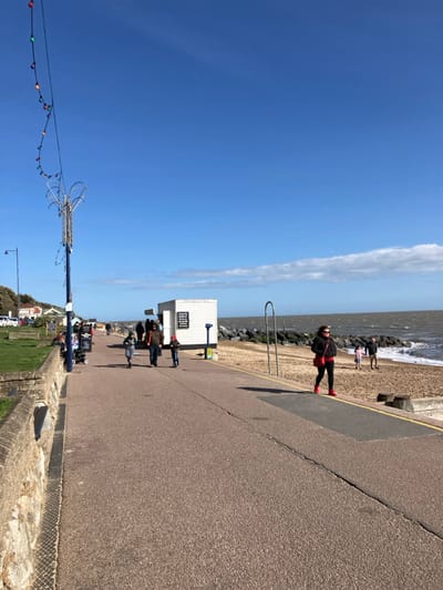 The image displays a sunlit coastal promenade in Felixstowe, United Kingdom. The sky is predominantly clear blue with a thin band of white clouds visible along the horizon. To the right, the grey-green sea features breaking waves and extends to the horizon, bordered by a shingle beach and a visible rock groyne.
The foreground is dominated by a wide, paved promenade of light brown aggregate, with a yellow line marking a section on the right. To the left, a low stone retaining wall separates the promenade from a grassy bank that ascends towards various buildings, including what appears to be a cafe, and parked vehicles. A tall blue lamppost with an ornate white top is positioned on the left, from which a string of multicolored fairy lights extends diagonally across the upper left portion of the frame.
Approximately ten individuals are present. In the mid-ground, a woman wearing a black jacket, red top, black trousers, and red shoes, carrying a red shoulder bag, walks from left to right on the promenade. Further along the promenade, an adult male walks with two children, both of whom are riding kick scooters. Behind them, another adult figure is visible. A white wooden kiosk, labeled "LONE SURVIVOR" with black signs attached, stands adjacent to the promenade on the beach side. Several people are congregated around this kiosk. On the beach, near the water's edge, two additional individuals (one adult, one child) are standing. A curved dark metal barrier, possibly a bicycle rack, is positioned on the promenade facing the beach.