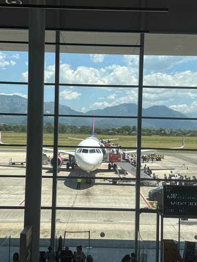The image depicts an airport tarmac scene, viewed from behind a large window with vertical and horizontal mullions. In the foreground, a large airplane, identified by its pink and white livery as likely a Wizz Air aircraft, is parked on the tarmac. Boarding stairs are attached to the front of the plane, with numerous passengers visible ascending and descending. A ground crew member in a yellow vest stands near the airplane's engine.
In the background, a mountainous landscape with a clear blue sky dotted with white clouds stretches across the horizon. Green fields and trees are visible between the tarmac and the mountains.
The airport environment is evidenced by the presence of the aircraft, tarmac markings, baggage carts, a service vehicle, and other airport equipment. People are seen in various states of boarding and disembarking the plane, indicating an active airport operation.
The question mentions the city of Kruja, Albania. While the image itself does not provide direct visual cues to confirm this location, the presence of a Wizz Air aircraft suggests a European context. If this scene is indeed in Kruja, Albania, it would indicate the presence of an airport serving that region. The overall scene is one of travel and transit within an airport setting, with a backdrop of natural mountain scenery.