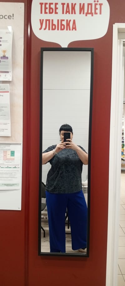 In Zhirnovsk, Russia, a woman is seen taking a mirror selfie in a retail environment. She is standing in front of a full-length mirror with a black frame. The mirror reflects her upper body and legs. She is wearing a dark, textured short-sleeved top and bright blue wide-leg pants. In her hands, she holds a black smartphone, angled to capture her reflection. Her hair is short and dark with bangs. She is wearing earrings and a wristwatch.

To the left of the mirror, a section of a wall displays various posters and informational sheets, including one with a golden trophy graphic and another with text in Cyrillic. The wall is painted a deep red color. To the right of the mirror, a white door frame is visible, and beyond that, shelves stocked with products can be seen, suggesting a commercial setting. The floor appears to be tiled. Above the mirror, a speech bubble graphic with red Cyrillic text is attached to the red wall.