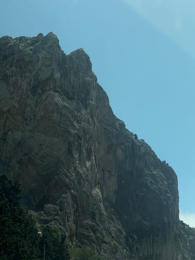 A low-angle shot depicts a mountain against a clear, light blue sky. The mountain occupies most of the frame, exhibiting a rough, rocky surface with various shades of gray. A few small trees cling to the mountainside. Patches of green foliage can be seen at the bottom right of the frame. The location is Орлиновский округ, Ukraine.