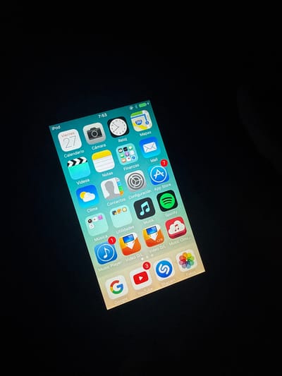 The media file presents an illuminated smartphone screen, likely an iPhone, tilted diagonally against a dark, featureless background. The screen displays the device's home interface, filled with rows of application icons and folders.

In the status bar at the top left, the time is visible as 7:53, alongside battery life, Wi-Fi, location services, and signal strength indicators.

The app icons are arranged in a grid. Several apps display notification badges: the 'Mail' app shows 7 unread notifications, 'App Store' has a badge (number unclear), 'YouTube' has 3, and a music app labeled 'Música' has 1.

Visible app icons and folder labels, predominantly in Spanish, include: 'Mapas' (Maps), 'Mail', 'App Store', 'Spotify', 'Video D/L', 'Music Clou...', 'Shazam', 'Fotos' (Photos), 'Reloj' (Clock), a folder labeled 'Finanzas' (Finance), a folder labeled 'Configuración' (Settings), 'Musik' (Music), a folder labeled 'Utilidades' (Utilities), 'Music Player', 'Video D/L', 'Google', 'YouTube', 'Cámara' (Camera), 'Notas' (Notes), 'Clima' (Weather), 'Contactos' (Contacts), 'Música' (Music). In the bottom row, there's a folder labeled 'iPod' and the 'Calendario' (Calendar) app, which displays 'Viernes 27' (Friday 27th) and 'Videos'. The dark background suggests the image was captured in low light conditions.