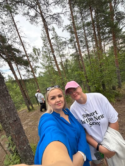 In the city of Kosalma, Russia, a selfie is taken in a wooded area with tall pine trees. Two women are the primary subjects. The woman in the foreground on the left is wearing a vibrant blue dress and has blonde hair. To her right, another woman is wearing a white t-shirt with the text "Smart Is the New Sexy" and a pink baseball cap. She is also wearing glasses. In the background, a man with a light-colored shirt and dark pants is walking away from the camera, seemingly on a dirt path bordered by a metal fence. The environment is characterized by dense forestation, with the sky visible through the tree canopy.