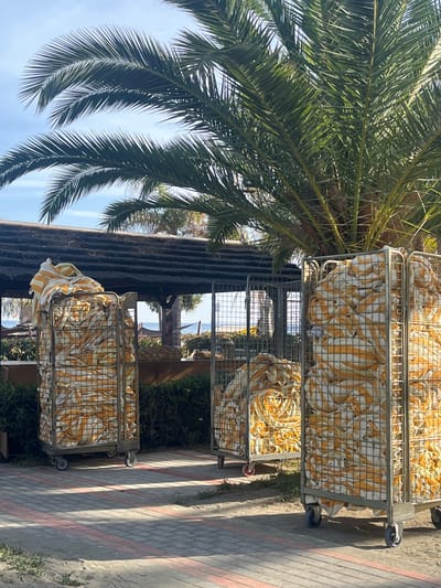 The image depicts three metal carts filled with folded yellow and white striped towels, positioned outdoors. A palm tree is visible to the left. A building with a thatched roof is behind the carts. The location appears to be in Germasogeia, Cyprus, given the architecture and flora. The ground is paved with gray bricks, marked with alternating red lines.