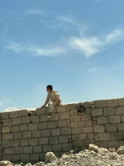 A young boy in a light-colored outfit sits on top of a low brick wall in Deh-E Bagh, Afghanistan. The wall is made of gray bricks, and there is debris and rubble at its base. The sky is light blue with some clouds. The boy is looking to the right of the frame.