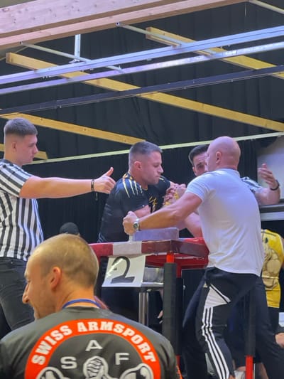 An indoor arm-wrestling competition is depicted in Reisenberg, Austria. The primary focus is an arm-wrestling match between two men at a red and brown table marked with the number "2". The man on the left wears a black shirt with yellow lightning patterns and intensely grips his opponent's hand. The man on the right, bald and wearing a white t-shirt and black Adidas track pants, also focuses on the match, gripping a table peg with his left hand. A referee, dressed in a black and white striped shirt, stands to the left, extending his left arm with a thumbs-up gesture. In the foreground, partially obscured, a man with light brown hair wears a black "SWISS ARMSPORT SAF" shirt, looking towards the match. The background features a dark fabric backdrop, with wooden beams and metal supports visible in the ceiling structure.