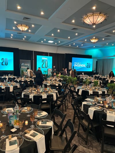 A conference room is set for an event, likely a banquet. Round tables with black tablecloths and transparent place settings are arranged in rows. Black folding chairs surround the tables. A stage with multiple screens displaying "Project Inspire 2.0" is visible at the front of the room. Several people are seated on the stage. The ceiling is decorated with ornate light fixtures and recessed lighting. The room appears to be well-lit.