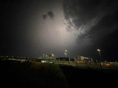 The image captures a nighttime scene at the intersection of the A9 and A4 highways near Badhoevedorp, Netherlands, during a thunderstorm. A bolt of lightning illuminates the dark, turbulent sky, casting an eerie glow on the surroundings. The highway stretches out, lined with streetlights that pierce through the gloom. Various road signs are visible, indicating directions and speed limits. The foreground shows a grassy embankment, hinting at the elevated position from which the photograph was taken. The atmosphere is charged with the raw power of nature, showcasing a dramatic interplay of light and shadow.