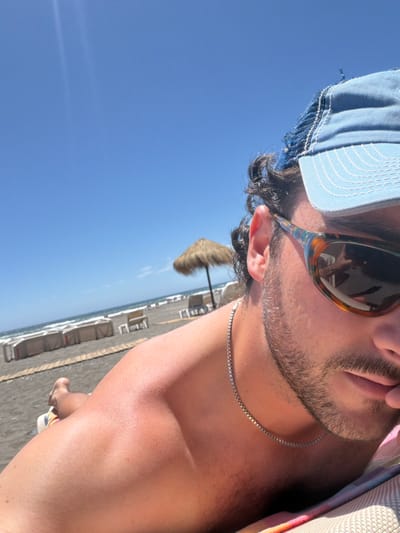 A person wearing sunglasses and a blue baseball cap is captured in a close-up shot, facing slightly away from the camera. A thin silver necklace adorns the neck. In the background, a beach scene unfolds under a bright blue sky. The beach is dotted with beach chairs and a straw umbrella, suggesting a sunny, relaxing environment.