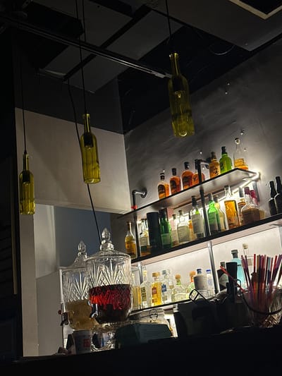 The image shows a dimly lit bar in Xuhui District, China. The bar features several shelves stocked with various bottles of alcohol. Hanging from the ceiling are three light fixtures made from repurposed green glass bottles. In the foreground, two large glass dispensers are visible, one filled with a red liquid and the other with a yellow liquid. A collection of colorful straws is in a glass container to the right. The overall ambiance is dark and atmospheric, with the shelves illuminated by backlighting. There are no people visible in the image.
