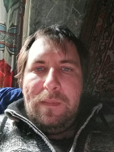 A self-portrait depicts a middle-aged man with fair skin, blue eyes, and short brown hair with bangs. He has a full beard and mustache. The man is wearing a gray and white patterned sweater with a dark lining around the collar.
The background includes a patterned curtain to the left, a textured wall or fabric directly behind him, and a dark patterned tapestry to the right. The lighting appears dim.
The location of Parabel, Russia is not discernible in the image.