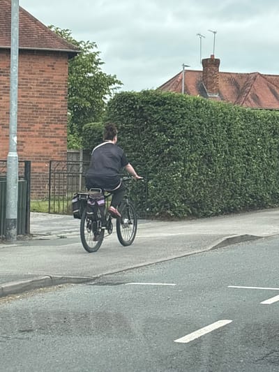 A person rides a bicycle with saddlebags on a paved sidewalk in Crewe, United Kingdom. The person is wearing a dark t-shirt and dark trousers, and their hair is dark and curly. The bicycle has a dark frame and black wheels. The background includes a brick building on the left, a dense green hedge in the center and right, and a residential roofline with chimneys and antennas in the distance. The sky is overcast. The road in the foreground has white markings.
