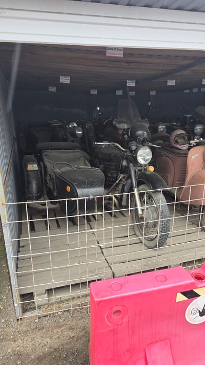 The image shows a collection of vintage motorcycles and scooters parked inside a metallic shed or garage. The motorcycles are various models, including one with a sidecar in the foreground. Several signs with Cyrillic text are visible on the wooden ceiling of the shed, identifying some of the vehicles and indicating they are "Made in USSR." The environment appears to be outdoors, with a gravel ground and a red plastic barrier in the foreground. The overall scene depicts a storage area for antique vehicles. The location is identified as Bol'shoi Mashlyak, Russia.