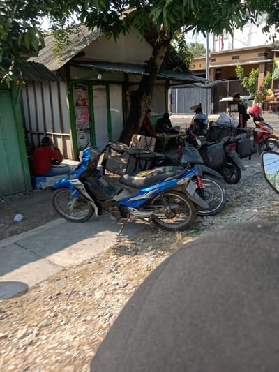 The image depicts an outdoor scene near a small building, possibly a shop or eatery, with motorcycles parked in front. The building has a corrugated metal roof and a green-painted frame around its entrance. Several people are seated at a table outside, appearing to be engaged in conversation or eating. A man in a red shirt is sitting on a blue box on the left side of the frame, facing away from the camera. Two motorcycles are prominently featured in the foreground, one blue and black, and another black and grey, both equipped with large bags or cargo carriers on the back. In the background, a red motorcycle is visible, as is a house with a fence and some trees. The ground is covered with gravel and dirt. The overall impression is of a casual gathering in a roadside setting in Glagah Wangi, Indonesia.