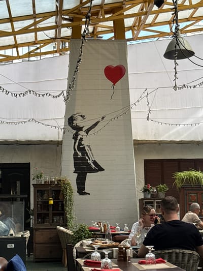 The image captures a bustling outdoor seating area of a restaurant in Zhukovka, Russia. A prominent mural of a girl releasing a red heart-shaped balloon adorns a white pillar, drawing the eye. String lights are draped across the ceiling and from the pillar, adding a festive ambiance. Several tables are set for dining with red napkins and glassware visible. In the foreground, a couple is seated at a table, with the woman wearing sunglasses and the man facing away from the camera. Further back, another man is seated alone, looking towards the right. The environment appears to be an open-air structure with a covered roof supported by wooden beams. A vintage wooden cabinet filled with jars and decorative items sits against a wall, adding to the restaurant's character. The overall scene suggests a lively and inviting dining experience.
