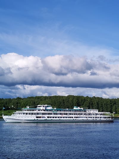 The image shows the river cruise ship "Ivan Kulibin" sailing on the Volga River. The ship is white with green trim and has several decks. The river is blue and there is a forested shoreline in the background. Above the shoreline are clouds. The image was taken in the city of Rybinsk, Russia.