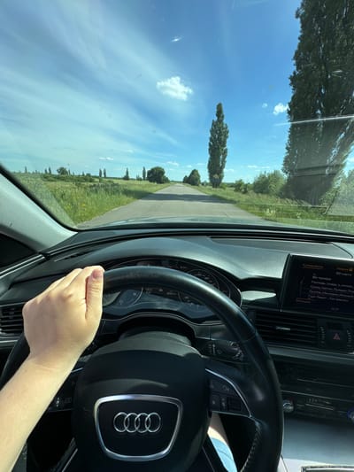 The image captures a first-person perspective from the driver's seat of a car, likely an Audi, as it travels along a paved road through a rural landscape. The driver's left hand is visible on the steering wheel, a portion of the dashboard with a digital display and climate controls are also in view. The road ahead is straight, bordered by lush green fields and tall, slender trees on either side. The sky above is a bright blue with scattered white clouds, indicating a sunny day. The digital display on the dashboard shows navigational information and media playback details, including song titles in Ukrainian. The scene suggests a journey through the countryside, possibly near the village of Dovhe, Ukraine, given the car's navigation system and the characteristic landscape.