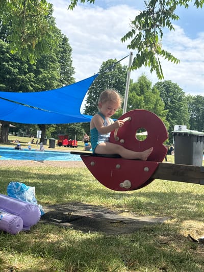 In Giessen, Germany, a young child with blonde hair, wearing a blue swimsuit, sits on a red playground apparatus with a circular cutout. Their legs are extended, and their right hand grasps a tan-colored bar. The child appears focused on an object in their hand. The playground structure is made of red plastic and wood, supported by a thick wooden beam.

In the background, a blue canopy stretches across part of the scene, casting a shadow. Beyond the canopy, a shallow blue pool is visible, with several people of various ages relaxing or playing in the water. Some children are seen near a red toy train. Trees with lush green foliage surround the area, indicating a pleasant outdoor setting, likely a park or a family-friendly recreational area. The sky above is blue with scattered white clouds. The lighting suggests it is daytime, with the sun casting shadows. Near the playground, some purple inflatable toys and a blue plastic bag lie on the grassy ground. The overall atmosphere appears relaxed and enjoyable, with families taking advantage of the warm weather for outdoor activities. No visible text is present in the image.