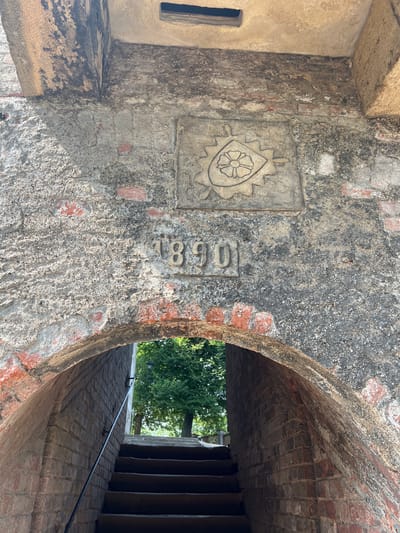 This is an eye-level, medium shot of an arched stone passageway. The rough, textured stone walls of the archway show signs of age and wear, with visible bricks peeking through the mortar. Above the arch, a relief carving of a stylized flower or rose within a shield is set into the stone, with the numbers "1890" carved below it. The passageway leads to a set of dark stairs, with a black metal handrail on the left side. The stairs ascend to an outdoor area where green trees and a lamppost are visible in the distance. The lighting suggests a sunny day, with shadows cast by the archway. This passageway is part of the Náchod Castle complex in Náchod, Czechia, likely an entrance or service area within the historical fortifications.