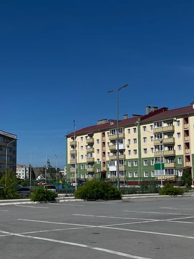 This is a photograph of a residential complex in the city of Achinsk, Russia, under a clear blue sky. The foreground features an empty asphalt parking lot marked with white lines. Beyond the parking lot, a green fence encloses a playground area with colorful equipment. Adjacent to the playground is a modern, multi-story apartment building with a red tiled roof. The building is painted in sections of light yellow and green. Numerous balconies with railings are visible along the facade. To the left of this building, another apartment building, also predominantly yellow, can be seen, along with streetlights and trees. In the background, other buildings and trees are visible, suggesting a suburban or urban residential area. The overall impression is one of a well-maintained, typical Soviet-era residential block.