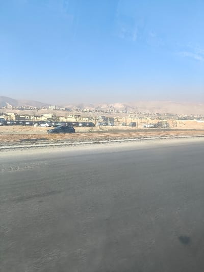 The image depicts an outdoor daytime scene in Dahiyat Harasta, Syria, offering a wide view of a semi-arid landscape. In the immediate foreground, a broad, dark grey asphalt road surface occupies the lower half of the frame. Beyond the road, a strip of light brown, dusty terrain transitions into an unpaved parking area containing approximately two dozen parked vehicles. These vehicles include multiple dark-colored sedans and SUVs, such as a prominent dark grey SUV in the central midground. A low, light-colored barrier or fence runs along the edge of the parking area. In the background, numerous light-colored, low-rise residential buildings with flat roofs are scattered across a rising, dry, and undeveloped hillside. The far background features dusty, light brown hills or mountains under a clear, pale blue sky with visible atmospheric haze. No individuals are discernible, and no active interactions or movements are depicted.