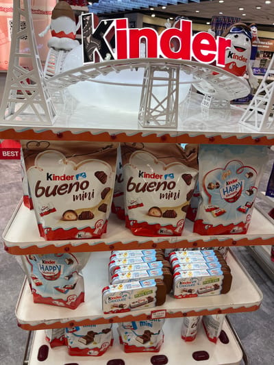 The image depicts a retail display of Kinder chocolate products, primarily Kinder Bueno Mini and Kinder Happy Moments, at an airport in Katunayake, Sri Lanka. The display features a prominent Kinder logo in bright red lettering and several decorative elements, including white scaffolding-like structures and white inflatable figures resembling Kinder characters. The products are arranged on tiered white shelves with orange trim. The background shows other retail displays and lighting fixtures typical of an airport concourse. The focus is on the various packaging of Kinder chocolates, with close-ups of the candy inside. The lighting is bright, suggesting an indoor commercial setting.
