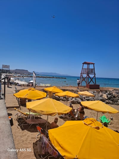 The image captures a vibrant beach scene on a clear, sunny day in Gouves Municipal Unit, Greece. The sky is a cloudless, deep blue, with a white airplane visible in the upper left quadrant, flying from left to right.

The horizon line separates the sky from the light blue-turquoise sea, which appears calm with minimal wave activity. Distant mountains stretch across the horizon. Rocky formations are present along the shoreline and extend into the sea. Several individuals are visible swimming or wading in the water.

On the sandy beach, a prominent feature is a reddish-brown wooden lifeguard tower with a small roof, positioned on a rocky outcrop near the water's edge on the right side of the frame. A person in a white shirt and dark shorts is depicted ascending the ladder of this tower.

The foreground and midground are dominated by numerous bright yellow beach umbrellas providing shade. Beneath these umbrellas, various sun loungers are arranged on the light brown sand; some are brown, others white, with towels draped over them. Several people are relaxing on or near these loungers; one individual with a bald head is seated facing away from the camera, while another person in a dark two-piece swimsuit stands near the loungers. A green inflatable alligator toy and a black and white soccer ball are visible on the sand towards the right.

Along the left side of the scene, a wooden boardwalk runs parallel to the beach, with a paved path adjacent to it. A white sign with black text, reading "minas grill & spiros," is mounted on a pole near the boardwalk, angled towards the beach. A person wearing a backpack stands on the wooden boardwalk, looking towards the beach. Further down the beach on the left, several white beach umbrellas are present, with more people gathered under them and near the water. A white banner with partial black text is also visible near the water's edge on this side.

The bottom-left corner of the image contains a white watermark stating "Galaxy A55 5G."