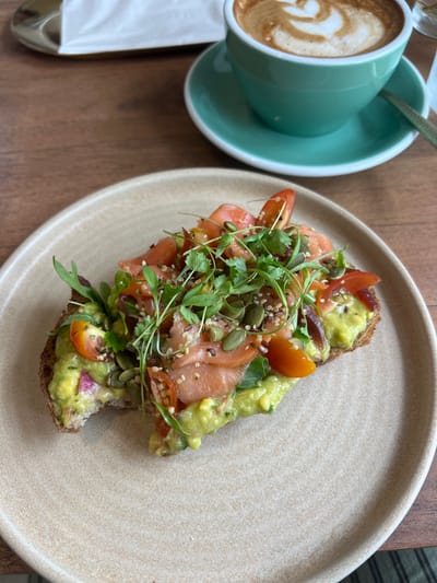 A partially eaten open-faced sandwich on a beige plate sits on a wooden table. The sandwich consists of a slice of toasted bread topped with smashed avocado, sliced tomatoes, smoked salmon, and fresh greens. There is a cup of coffee in the background. The scene is set in a cafe in Bella Vista, Panama, located at latitude 8.98 and longitude -79.519.