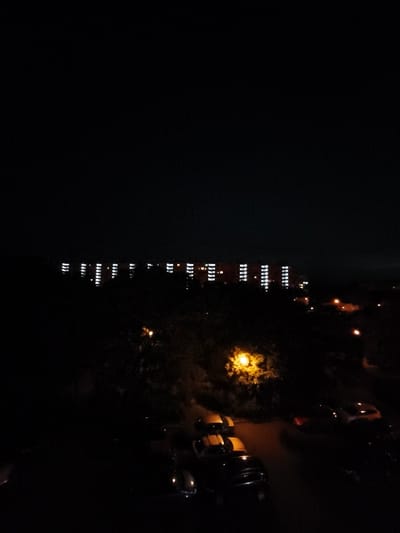 The image captures a nighttime scene in the city of Most, Czechia, characterized by the dark sky and the illumination from buildings and streetlights. In the background, a long, low-rise building complex is visible, with numerous windows lit up, creating a pattern of bright rectangles against the darkness. The foreground reveals a residential area, with trees and parked cars. Several vehicles are visible, some with their headlights or taillights illuminated. A distinct streetlamp with a yellow glow is prominent, illuminating the surrounding foliage. The overall atmosphere is one of a quiet night in a city environment.