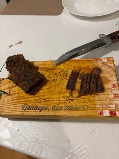 The image depicts a cutting board with biltong and a knife on a table. The cutting board has red and white markings on its top edge and writing on the side. Several sliced pieces of biltong are visible on the board, as well as a larger, uncut piece tied with green string. A knife with a wooden handle is positioned on the left side of the board, near the sliced biltong. A white plate is partially visible in the upper left corner of the image. The table has a textured white surface. The location is not identifiable as Lelystad, Netherlands from the provided image.